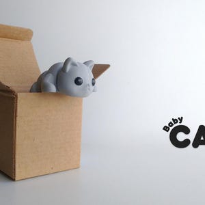 May include: A grey 3D printed baby cat figurine peeking out of a brown cardboard box. The text "Baby CAT" is printed in black on a white background.