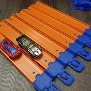 K&ouml;nnte beinhalten: Zwei Spielzeugautos, eines rot und eines schwarz, fahren auf einer orangefarbenen Rennstrecke mit blauen Verbindungsst&uuml;cken. Die Strecke besteht aus mehreren Teilen, die zusammengef&uuml;gt werden.