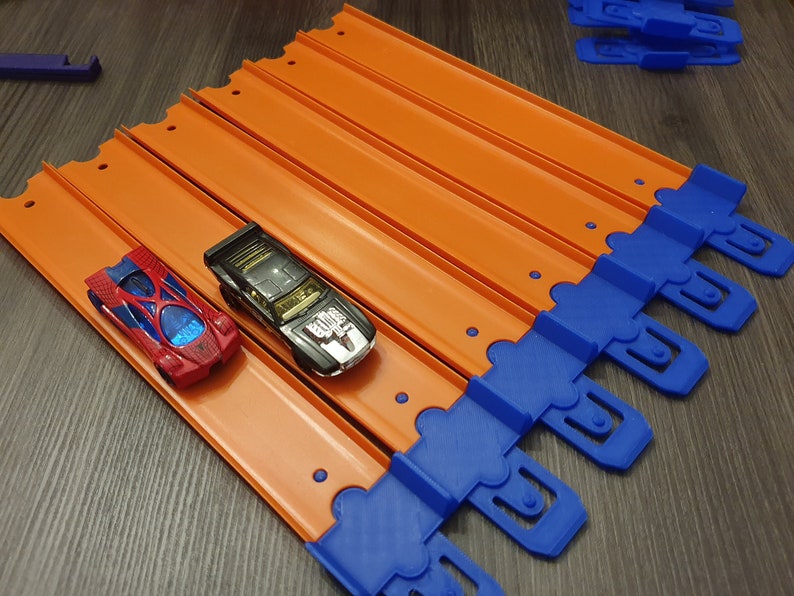 K&ouml;nnte beinhalten: Zwei Spielzeugautos, eines rot und eines schwarz, fahren auf einer orangefarbenen Rennstrecke mit blauen Verbindungsst&uuml;cken. Die Strecke besteht aus mehreren Teilen, die zusammengef&uuml;gt werden.