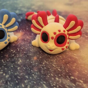 May include: Two 3D printed axolotl figurines. One is white with red accents and black eyes, the other is blue with white accents and black eyes.
