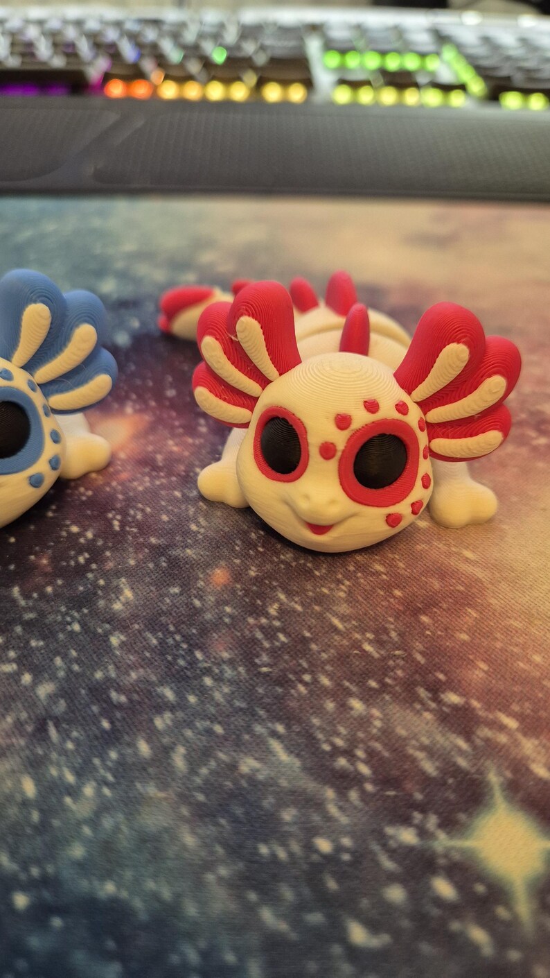 May include: Two 3D printed axolotl figurines. One is white with red accents and black eyes, the other is blue with white accents and black eyes.