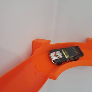 May include: A black toy car with a silver engine detail is driving on an orange plastic track. The track is curved and goes up a wall.