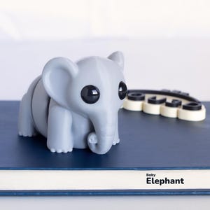 Op de afbeelding: Een grijze, gesegmenteerde olifantenfiguur met grote zwarte ogen en een gekrulde slurf staat op een blauw oppervlak. De tekst "Baby Elephant" staat onder de figuur. Een zwart-wit object staat achter de olifant.