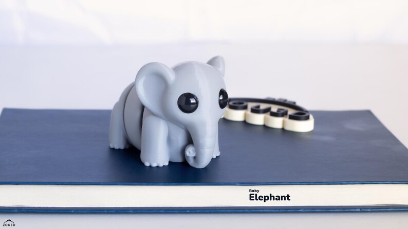 Op de afbeelding: Een grijze, gesegmenteerde olifantenfiguur met grote zwarte ogen en een gekrulde slurf staat op een blauw oppervlak. De tekst "Baby Elephant" staat onder de figuur. Een zwart-wit object staat achter de olifant.
