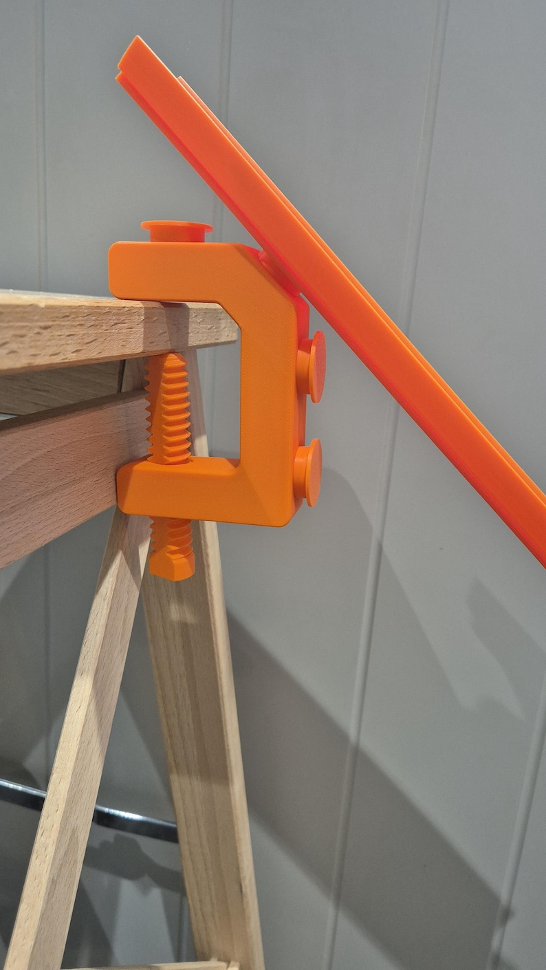 May include: An orange clamp attached to a wooden surface. The clamp is holding a long, orange piece of plastic.