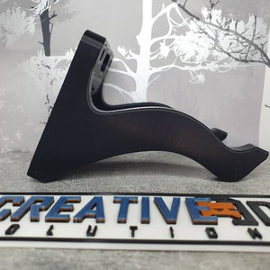 May include: A black plastic wall mount with a curved design. The mount is designed to hold a device, such as a phone or tablet, in a horizontal position.