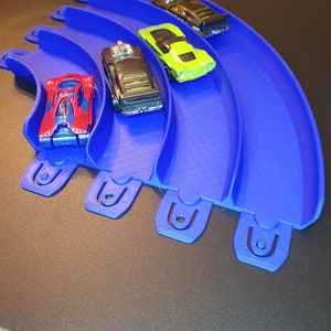 May include: A blue plastic track with five lanes for toy cars. Four toy cars are on the track, including a red car, a black car, a green car, and a black car.