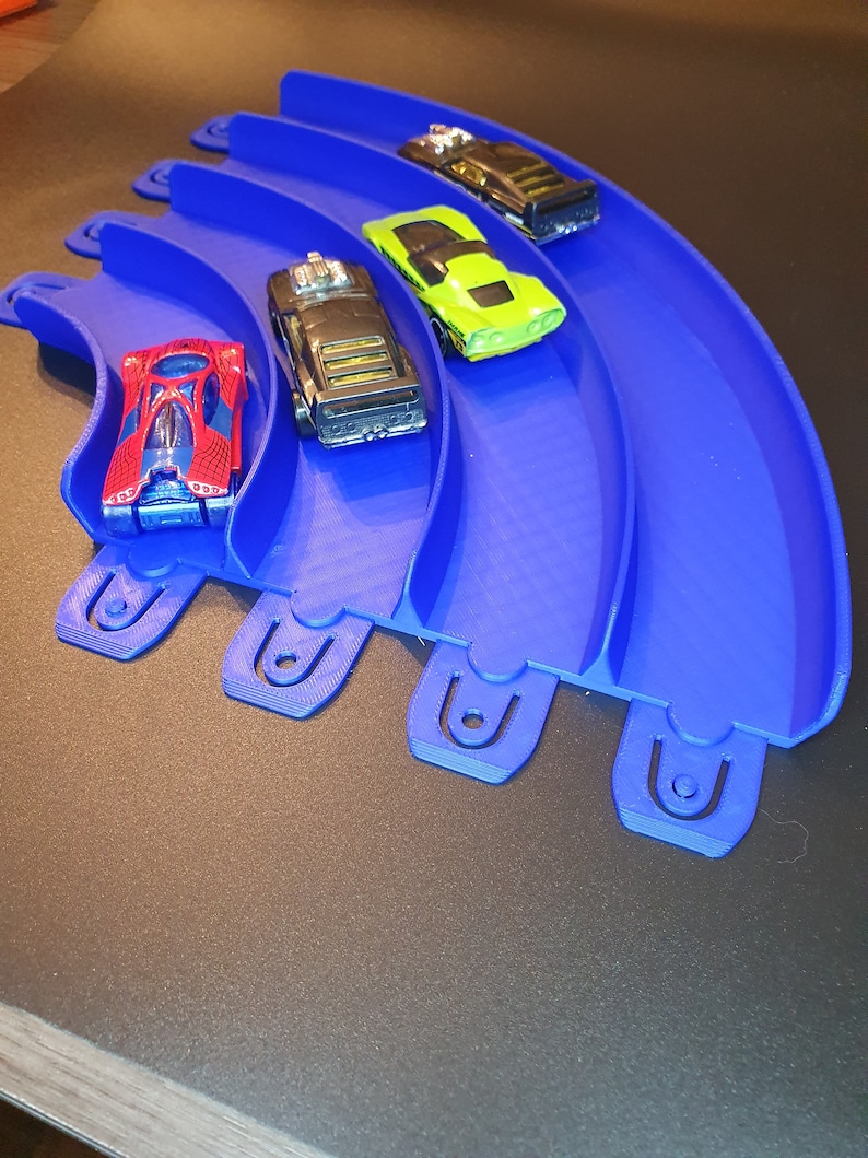 May include: A blue plastic track with five lanes for toy cars. Four toy cars are on the track, including a red car, a black car, a green car, and a black car.