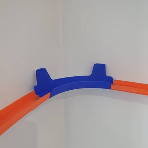 May include: A blue plastic track piece for a toy car race track. The track piece is designed to fit in a corner and has two orange track pieces attached to it.