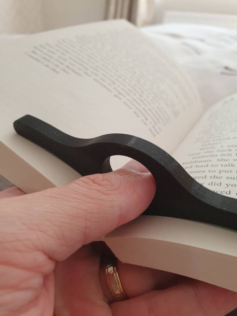 May include: A black plastic page holder for books. The page holder has a curved design and a hole for a finger to hold it in place.