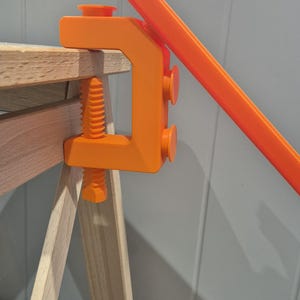 Op de afbeelding: Een oranje klem bevestigd aan een houten oppervlak. De klem houdt een lang, oranje stuk plastic vast.