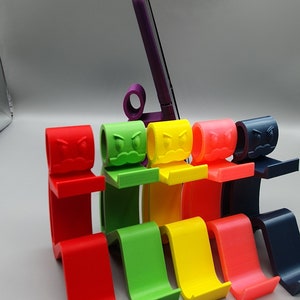 May include: Six colorful phone stands with a cartoon angry face design. The stands are made of 3D printed plastic and are arranged in a row from left to right: red, green, yellow, orange, blue, and black.