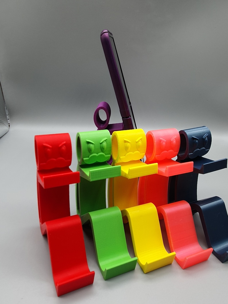 May include: Six colorful phone stands with a cartoon angry face design. The stands are made of 3D printed plastic and are arranged in a row from left to right: red, green, yellow, orange, blue, and black.