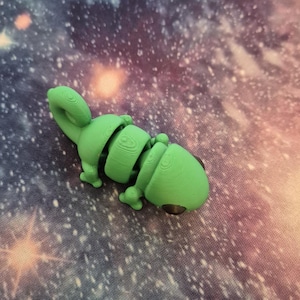 Op de afbeelding: Een groen, gearticuleerd kameleon beeldje met zwarte ogen. Het beeldje is geposeerd met zijn staart gekruld en zijn hoofd gekanteld naar de zijkant.