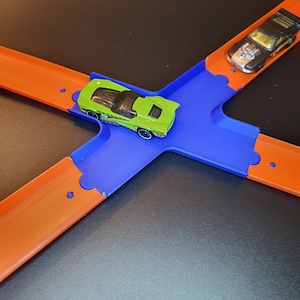 Op de afbeelding: Een groene speelgoedauto op een blauwe en oranje racebaanset. De auto bevindt zich op een blauw gedeelte van de racebaan, en de racebaan is verbonden met oranje gedeeltes.
