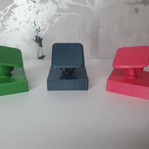 May include: Three 3D printed phone stands in different colours: green, blue, and pink. Each stand has a square base and a square top with a rounded edge. The stands are designed to hold a phone upright.