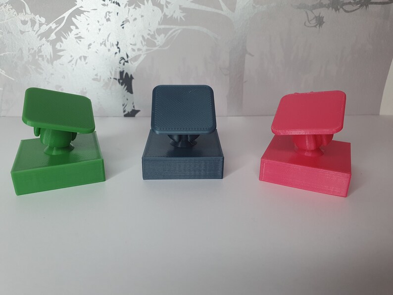 May include: Three 3D printed phone stands in different colours: green, blue, and pink. Each stand has a square base and a square top with a rounded edge. The stands are designed to hold a phone upright.