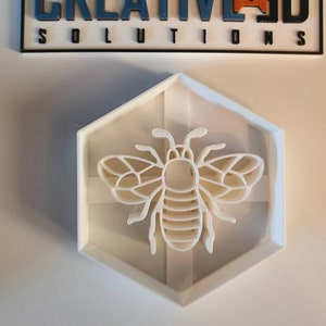 May include: White plastic hexagonal mold with a detailed bee design. The bee is in the center of the mold and has a textured body and wings.