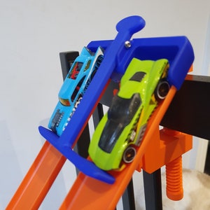 Op de afbeelding: Twee speelgoedauto's op een oranje baan, een blauwe en een groene, worden op hun plaats gehouden door een blauwe klem. De klem is bevestigd aan een zwarte houten constructie.