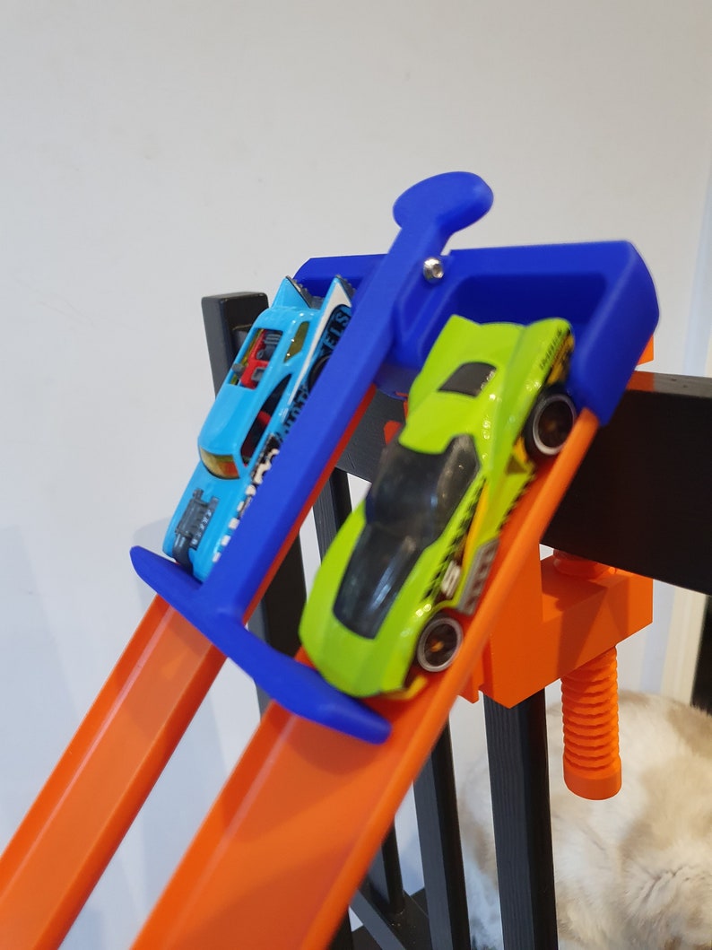 Op de afbeelding: Twee speelgoedauto's op een oranje baan, een blauwe en een groene, worden op hun plaats gehouden door een blauwe klem. De klem is bevestigd aan een zwarte houten constructie.
