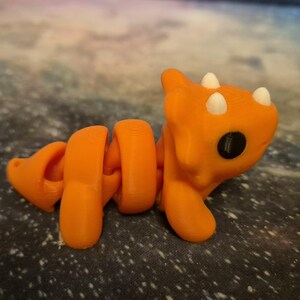 May include: Orange and white segmented toy dinosaur with a curled tail and three horns on its head.