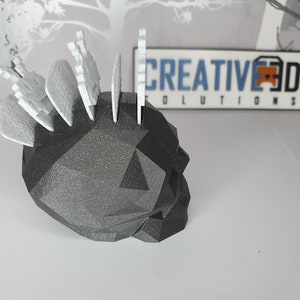 Puede incluir: Un soporte para p&uacute;as de guitarra en forma de calavera negra con un dise&ntilde;o geom&eacute;trico. El soporte tiene una fila de p&uacute;as blancas insertadas en la parte superior.