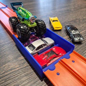 Puede incluir: Un cami&oacute;n monstruo Hot Wheels con dise&ntilde;o verde y azul, llamado "Carbonator", est&aacute; sobre una pista de carreras azul y naranja. Tambi&eacute;n se ven otros tres coches de juguete: un cup&eacute; amarillo, un sed&aacute;n plateado y un coche deportivo rojo.
