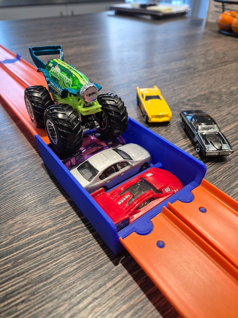Puede incluir: Un cami&oacute;n monstruo Hot Wheels con dise&ntilde;o verde y azul, llamado "Carbonator", est&aacute; sobre una pista de carreras azul y naranja. Tambi&eacute;n se ven otros tres coches de juguete: un cup&eacute; amarillo, un sed&aacute;n plateado y un coche deportivo rojo.