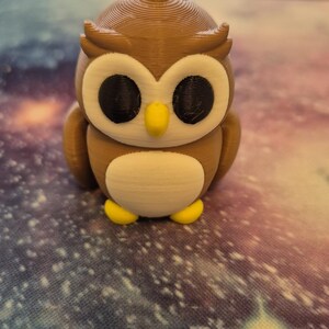 May include: A brown and white owl figurine with black eyes and a yellow beak. The owl is standing on a colorful surface.