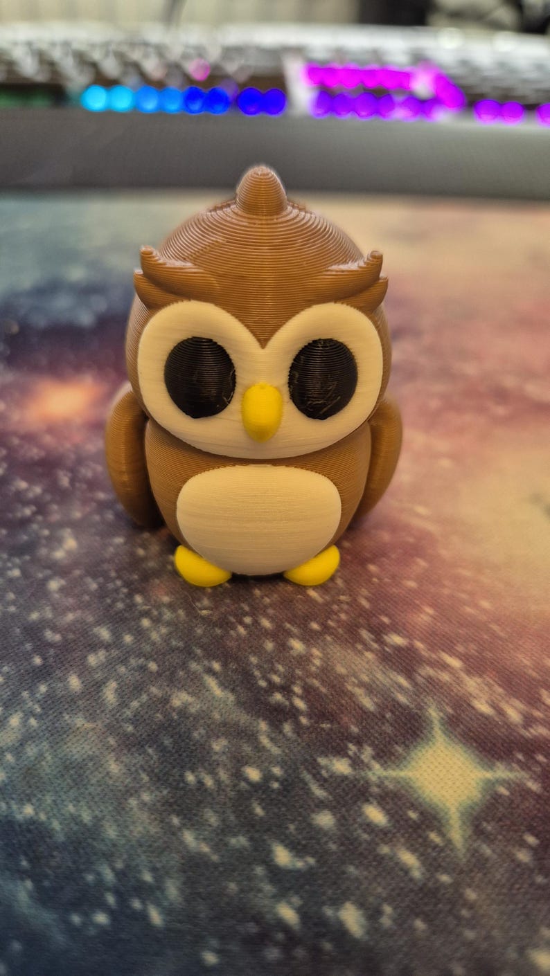 May include: A brown and white owl figurine with black eyes and a yellow beak. The owl is standing on a colorful surface.