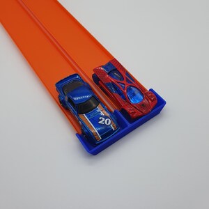 May include: Two toy race cars, one blue with the number 20 and orange accents, and one red with a web design, are positioned on a blue starting gate. They are on an orange race track, angled diagonally across a white background.