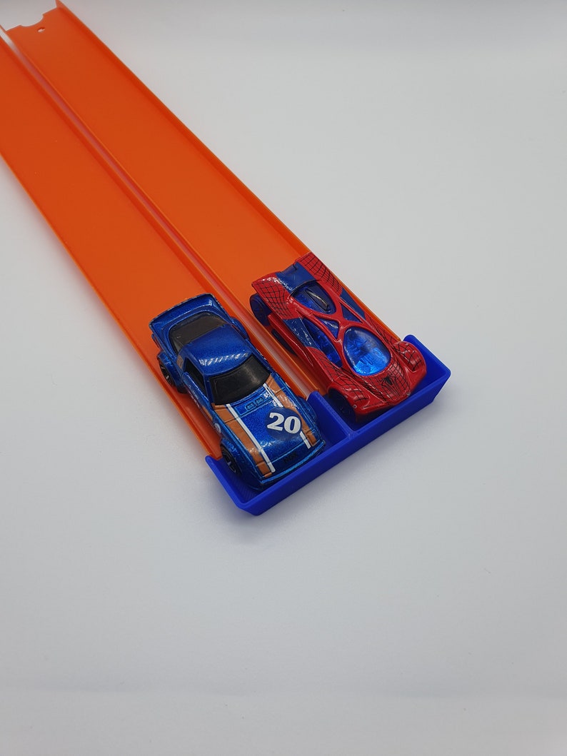 May include: Two toy race cars, one blue with the number 20 and orange accents, and one red with a web design, are positioned on a blue starting gate. They are on an orange race track, angled diagonally across a white background.