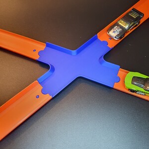 Op de afbeelding: Een blauwe plastic baan kruising voor een speelgoed auto racebaan. Twee speelgoedauto's zijn op de baan, een zwart en een groen.