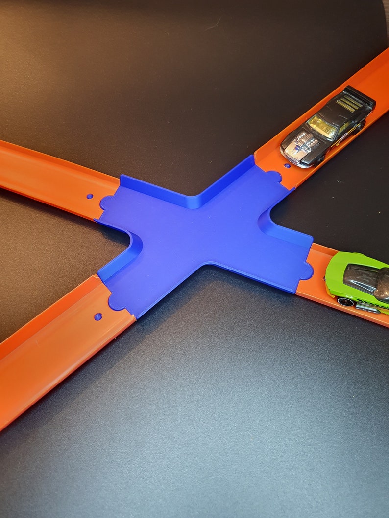 Op de afbeelding: Een blauwe plastic baan kruising voor een speelgoed auto racebaan. Twee speelgoedauto's zijn op de baan, een zwart en een groen.