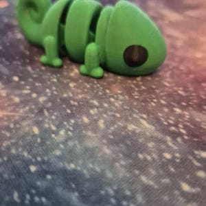 Op de afbeelding: Een groen plastic kameleon speelgoed met een gesegmenteerd lichaam en grote zwarte ogen. Het speelgoed ligt op een blauw-wit gevlekt oppervlak.