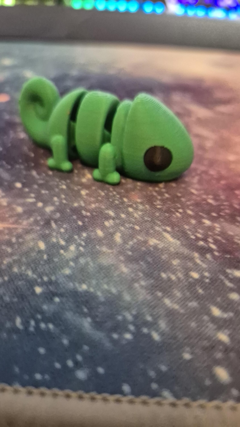 Op de afbeelding: Een groen plastic kameleon speelgoed met een gesegmenteerd lichaam en grote zwarte ogen. Het speelgoed ligt op een blauw-wit gevlekt oppervlak.