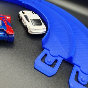 May include: A blue plastic race track with two toy cars. One car is red and blue with a spider web design. The other car is white with a black stripe and black wheels.