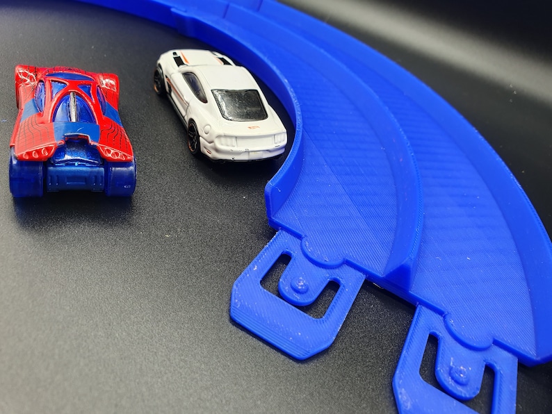 May include: A blue plastic race track with two toy cars. One car is red and blue with a spider web design. The other car is white with a black stripe and black wheels.
