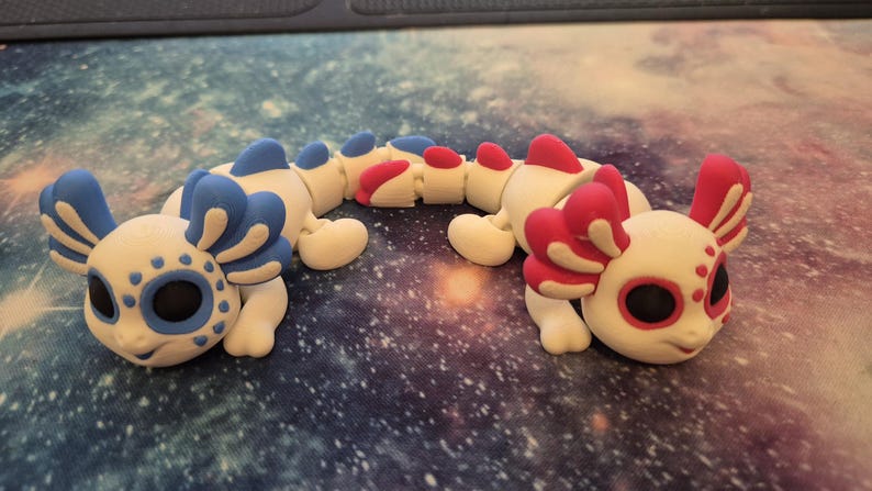 May include: Two white axolotl figurines with articulated bodies. One axolotl has blue and white gills and the other has pink and white gills.