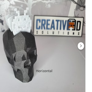 Puede incluir: Un macetero de calavera impreso en 3D negro y gris con una nave espacial extraterrestre impresa en 3D blanca en la parte superior. El macetero de calavera est&aacute; sobre una superficie gris. El texto "CREATIVE 3D SOLUTIONS" es visible en el fondo.