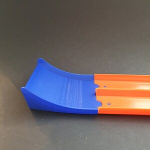 May include: A blue and orange plastic track piece for a toy car racing set. The blue section is a curved ramp and the orange section is a straight track.