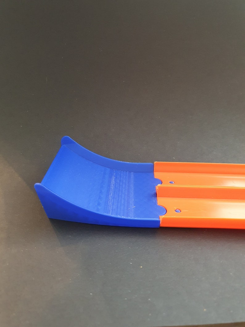 May include: A blue and orange plastic track piece for a toy car racing set. The blue section is a curved ramp and the orange section is a straight track.