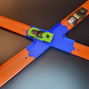 Op de afbeelding: Een speelgoedautobaanset met twee auto's, een groene en een zwarte, op een blauw en oranje baan.