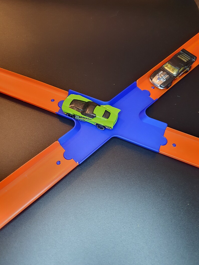 Op de afbeelding: Een speelgoedautobaanset met twee auto's, een groene en een zwarte, op een blauw en oranje baan.