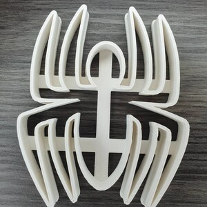 May include: White plastic spider cookie cutter. The cutter has a detailed design with multiple layers of lines and curves.