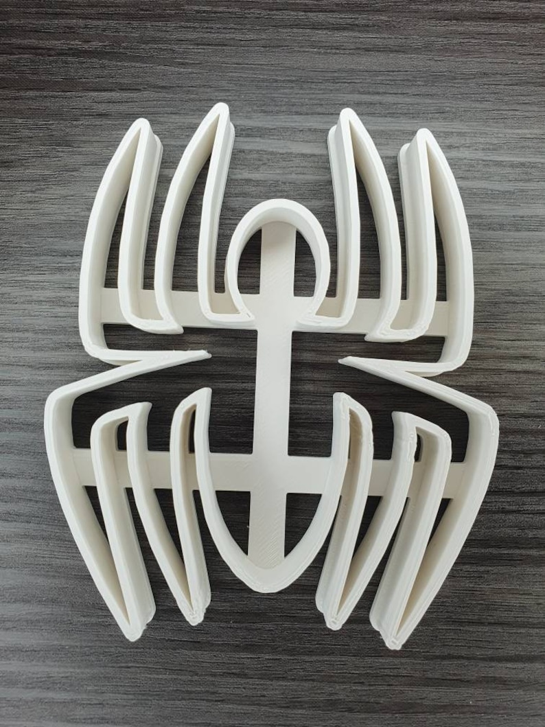 May include: White plastic spider cookie cutter. The cutter has a detailed design with multiple layers of lines and curves.