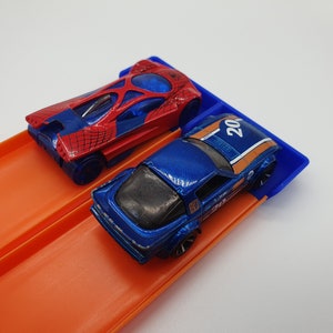 May include: Two toy race cars on an orange track. One car is red and blue with a spiderweb design, the other is blue with orange stripes and the number 20. The track has a blue starting gate.