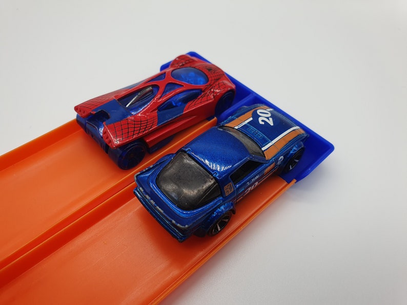 May include: Two toy race cars on an orange track. One car is red and blue with a spiderweb design, the other is blue with orange stripes and the number 20. The track has a blue starting gate.