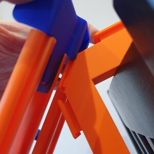 May include: A close-up of a blue and orange toy track set. The blue piece is a connector that fits into the orange track.