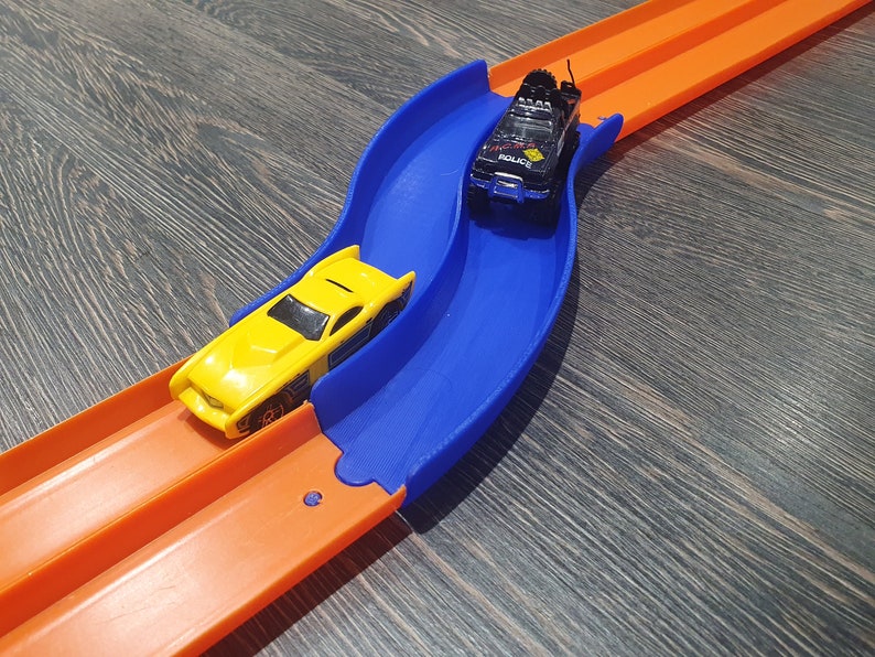 May include: A blue and orange plastic toy race track with two toy cars. One car is yellow and the other is black with the word "Police" on the side.